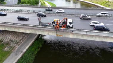 Köprü kenarında turuncu güvenlik donanımlı işçilerle bir inşaat işi. Yakınlarda bir kamyon var. Bölge kentsel altyapıyı gösteriyor.