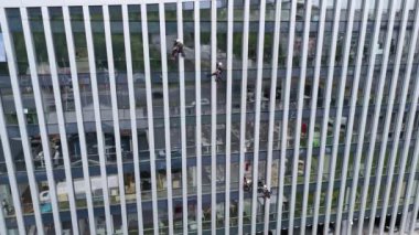 Modern bir cam binanın camlarını temizleyen işçiler, ipe asılmış. Yansıtıcı yüzeylere sahip yüksek katlı bir yapı gösteriliyor