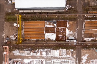 Metal kirişleri ve boruları olan bir inşaat alanının havadan görüntüsü. Zemin kısmen karla kaplı ve çeşitli malzemeler var. Sarı bir vinç görünüyor..