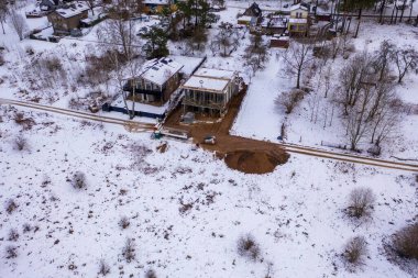 Karla çevrili bir inşaat alanının havadan görüntüsü. Sitede kısmen tamamlanmış, düz çatısı, toprak yolları ve karlı bir manzarası olan bir bina yer alıyor. Çevrede ağaçlar görülebiliyor, bu da kırsal bir alanı işaret ediyor..