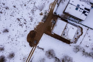 Karla çevrili bir inşaat alanının havadan görüntüsü. Sitede kısmen tamamlanmış, düz çatısı, toprak yolları ve karlı bir manzarası olan bir bina yer alıyor. Çevrede ağaçlar görülebiliyor, bu da kırsal bir alanı işaret ediyor..