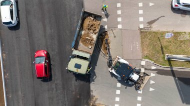 Ağır makineler ve işçilerle dolu bir yol şantiyesinin havadan görüntüsü. Ekipman büyük bir kazıcı, küçük bir yükleyici ve çeşitli aletleri içerir. Yol yüzeyi tamir ediliyor..
