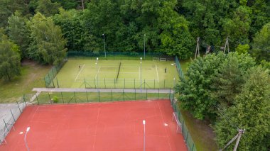 Tenis kortu ve yemyeşil ağaçlarla çevrili kırmızı çoklu spor kortunun yer aldığı bir spor kompleksinin havadan görünüşü. Oyuncular tenis kortlarında aktif bir açık hava ortamı sergileyerek görülürler..