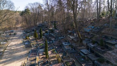 Uzun ağaçlarla çevrili bir mezarlığın havadan görünüşü. Mezarlar düzenli olarak düzenlenir. Manzara yeşillik ve mezar işaretlerinin karışımını gösteriyor, dingin bir atmosfer yaratıyor..