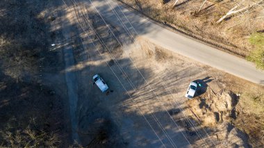 Toprak bir yolda park etmiş iki aracı gösteren kırsal bir bölgenin havadan görüntüsü. Çevresindeki manzara çoğunlukla çıplak ve yerde uzun gölgeler bırakan bazı ağaçlar var..