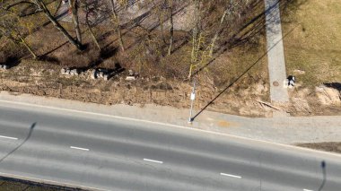 Yol kenarındaki bir hava manzarası dağınık tuğlaları ve kiri olan hasarlı bir kaldırımı gösteriyor. Çimenler kaldırım boyunca görünür, ve yakınlardaki ağaçların gölgeleri yere dökülür..