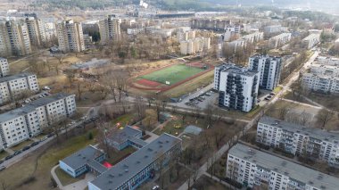 Yeşil alanları ve otoparkları olan konut ve ticari binaların karışımının yer aldığı modern şehir manzarası. Ufuk çizgisi çeşitli yapıları içerir.