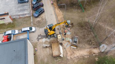 Konut binalarının yakınındaki bir inşaat alanının havadan görüntüsü. Ağır makineler kirli bir alana park edilmiş ve işçiler görülebilir durumda. Yakınlardaki bir otoparkta birkaç araba sıraya dizildi..