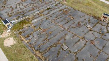 Çatlakları ve aşırı gelişmiş bitkileri olan terk edilmiş beton bir yüzey. Eskiden basketbol sahasıydı. Bölgede ihmalkarlık belirtileri var. Çimenler çatlaklardan büyüyor..