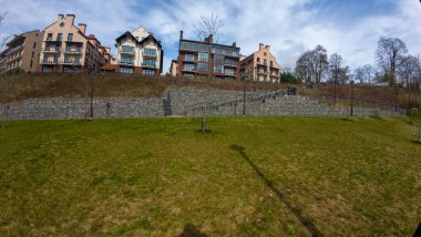 Modern binalara uzanan taştan bir merdiveni olan çimenli bir bölgenin manzarası. Binalarda, balkonları ve geniş pencereleri olan mimari tarzların bir karışımı yer alıyor. Gökyüzü parçalı bulutlu, dingin atmosfere ekleniyor.