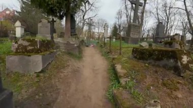 Mezar taşları ve demir çitlerle dolu eski bir mezarlığın içinden geçen bir kaldırım taşı yolu. Arka planda yaprakları olmayan ağaçlar ve mavi çiçekler görülebilir..