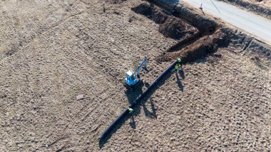 Aerial view of construction workers and an excavator installing a pipeline in a field. clipart