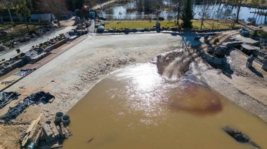 Göl kenarındaki bir inşaat alanının havadan görüntüsü. Bölge, çamurlu su, çakıl yolları ve peyzaj elementleri içeren toprak çalışmalarının bir karışımını gösteriyor..
