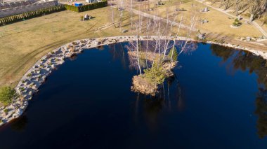 Bereketli bir yeşillik ve dolambaçlı bir patikayla çevrili sakin bir göletin havadan görünüşü. Gölet ağaçlarla kaplı küçük bir adaya sahiptir ve arazi kayalar ve otlarla beneklenmiştir..