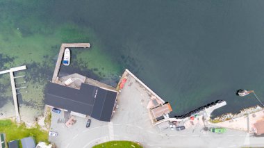 Limanı, park edilmiş arabaları ve konut binaları olan küçük bir sahil köyünün havadan görünüşü. Limanda küçük bir rıhtım ve bir tekne bulunurken, çevre bölgede yollar ve yeşillikler yer alıyor..