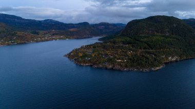 Ağaçlar ve birkaç evle çevrili sakin bir mavi fiyorda uzanan bir orman manzarası. Manzara berrak bir gökyüzünün altında yeşil ve sonbahar renklerinin karışımı ile huzurlu..