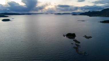 Küçük kayalık adaları ve bulutlu gökyüzünün altındaki uzak dağlarla sakin bir fiyordun havadan görünüşü.