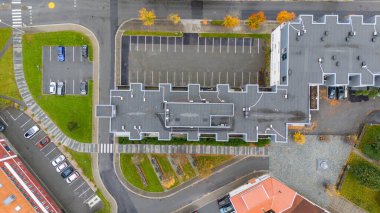 Yeşil alanlar ve yollarla çevrili, eşsiz bir çatı tasarımına sahip modern bir binaya yakın bir otoparkın havadan görüntüsü. Görüntü park halindeki arabaları ve sonbahar renkli ağaçları yakalıyor..
