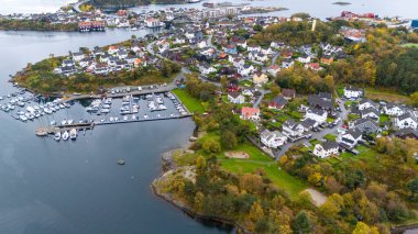 Evleri, marinaları ve yemyeşil yeşillikleri olan kıyı kasabasının havadan görünüşü. Manzarada limana yanaşmış, ağaçlar ve tepelerle çevrili yerleşim alanları ve tekneler yer alıyor..