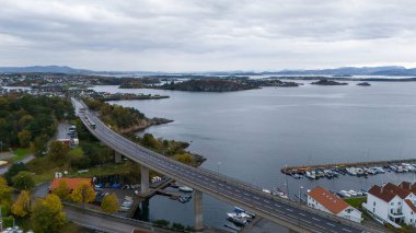 Suyun üzerinde köprüsü olan, adalarla çevrili ve yat limanına yanaşmış bir sahil kasabasının havadan görüntüsü. Sahne bulutlu bir gökyüzü ile kaplıdır ve sonbahar yaprakları görünür..