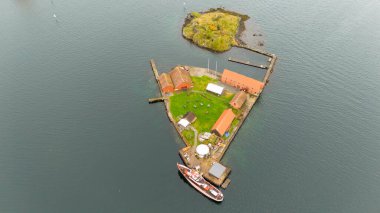 Suyla çevrili kırmızı çatılı küçük üçgen bir adanın havadan görünüşü. Adanın kenarına bir tekne yanaştı ve daha küçük bir ada yakınlarda görünüyor. Sahne huzur dolu..