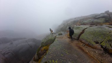 Sisli havada, yosun kaplı kayalar ve sınırlı görüş mesafesi olan kayalık bir dağ yolunda yürüyen iki yürüyüşçü..