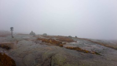 Islak kayalık arazi ve işaret direkleriyle sisli dağ zirvesi.