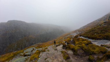 Aşağıdaki gizli vadiye bakan kayalık arazi ve seyrek bitki örtüsüyle sisli bir dağ manzarası. Yalnız bir yürüyüşçü, puslu sahneye bir ölçek duygusu ekleyerek kenarda durur..