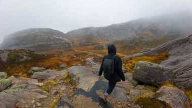 Siyah ceketli ve sırt çantalı bir adam sisli bir dağ manzarasında kayalık bir patikada yürüyüş yapıyor..