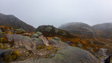 Siyah ceketli ve sırt çantalı bir adam sisli bir dağ manzarasında kayalık bir patikada yürüyüş yapıyor..