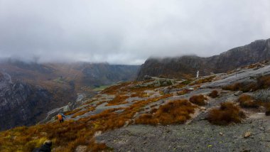 Bir yürüyüşçü bulutlu bir gökyüzünün altında engebeli arazi ve seyrek bitki örtüsüyle çevrili kayalık bir dağ yolu boyunca yürür..