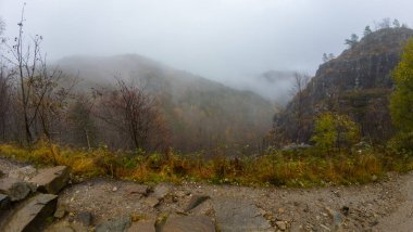 Puslu bir dağ manzarası. Tepeleri kaplayan bir sis ve ön planda kayalık bir yol..