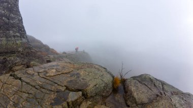 Kayalık arazide yürüyüşçülerle sisli dağ manzarası.