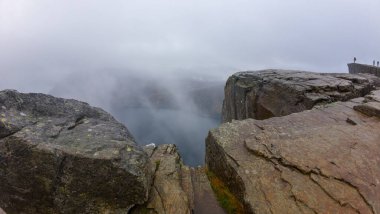 Bir su kütlesine tepeden bakan kayalık bir kayanın olduğu sisli bir manzara, uzak görüşü sisli bir şekilde gizliyor. Sahne sakin ve atmosferik. Uçurumun kenarında birkaç kişi var..