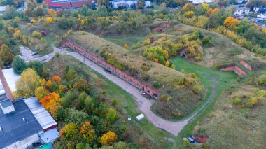 Sonbahar yeşillikleriyle çevrili eski bir kalenin havadan görünüşü, kemerli kapıları ve çimenli çatıları olan uzun tuğla bir yapı içeriyor. Manzara sonbahar renklerinde ağaçlarla benekli..