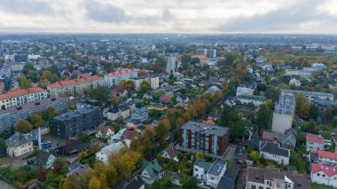 Konut binaları, yeşil ağaçlar ve sonbahar yapraklarıyla dolu bir banliyö bölgesinin havadan görünüşü. Ufuk çizgisinde alçak binalar ve arka planda bulutlu bir gökyüzü yer alıyor..