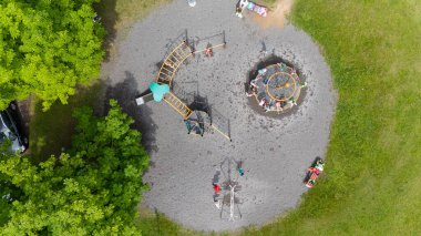 Çimlerle ve ağaçlarla çevrili, ekipmanlarla oynayan çocukların olduğu bir oyun parkı..