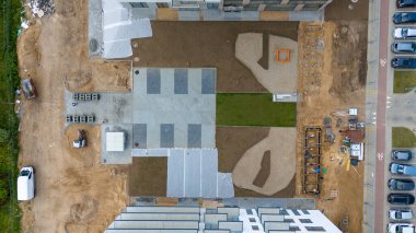 Kısmen tamamlanmış avlusu, asfaltlı alanları, çimen bantları ve çevre binaları olan bir inşaat alanının havadan görüntüsü..