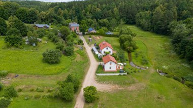 Birkaç ev, toprak yol ve yemyeşil yemyeşil verimli bir arazinin havadan görünüşü. Sahne ağaçlar, bahçeler ve açık alanlardan oluşuyor, sakin bir kırsal atmosfer yaratıyor..