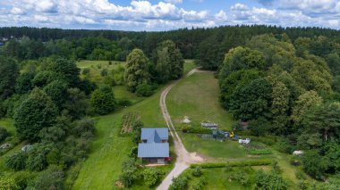 Yeşillik ve parçalı bulutlu gökyüzünün altındaki ağaçlarla çevrili, küçük bir evi olan kırsal bir arazinin havadan görünüşü..