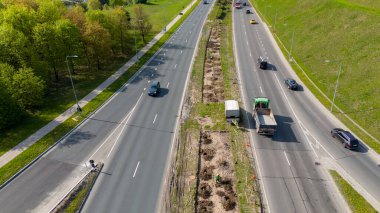 Bölünmüş bir otoyolun her iki yönde de iki şeritli havadan görünüşü, işçilerin ağaç diktiği merkezi çimenli bir medyanı içeriyor. Arabalar yolda görünüyor ve çevresi de yeşil..