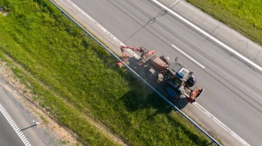 Çim biçme makinesiyle yol kenarındaki çimenleri kesen traktörün hava görüntüsü.