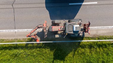 Çim biçme makinesiyle yol kenarındaki çimenleri kesen traktörün hava görüntüsü.
