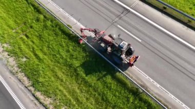 Çim biçme makinesiyle yol kenarındaki çimenleri kesen traktörün hava görüntüsü.