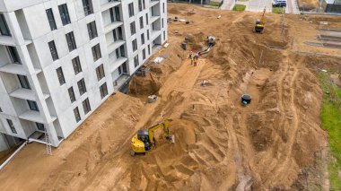 Ağır makinelerin, işçilerin ve kazılmış toprağın geniş bir alanının bulunduğu bir inşaat alanının havadan görüntüsü. Bölge kısmen inşa edilmiş bir bina ve çeşitli malzemelerle çevrili..