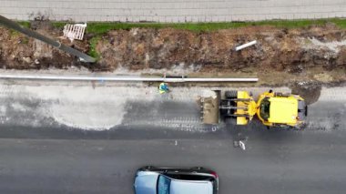 Yol üzerinde çalışan sarı kazıcılı bir inşaat alanının havadan görüntüsü. Bir işçi kaldırım inşa ediyor..