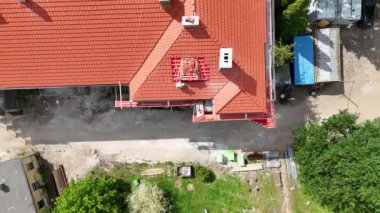 Ağaçlar ve inşaat malzemeleriyle çevrili kırmızı kiremitli bir çatıdaki inşaat işçisinin hava görüntüsü. Olay yeri, kısmen tamamlanmış çatısı ve yakınlarındaki yapıları olan bir yerleşim alanı yakalıyor..