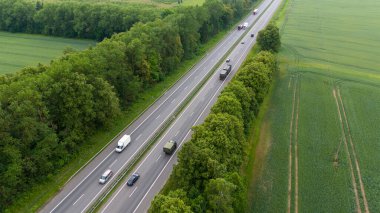 Yeşillik yeşil tarlalar ve ormanlar boyunca uzanan bir otoyolun havadan görünüşü, her iki yönde de çok sayıda araç var. Sahne, doğa ve altyapı arasındaki zıtlığı yakalıyor..