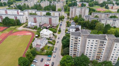 Apartman binaları, spor sahaları ve cadde boyunca park edilmiş arabaların bulunduğu bir yerleşim yeri manzarası. Sahne yeşil ağaçlarla dolu ve şehir altyapısı ile çevrili..