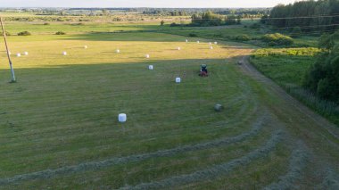 Dağınık saman balyalarıyla geniş bir alanda çalışan bir traktörün hava görüntüsü..